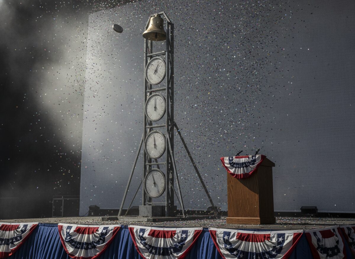 Press photo: a carnival high-striker strength-test game on a bunting-draped stage, the puck rocketing toward the bell, blank gauge dials mounted alongside